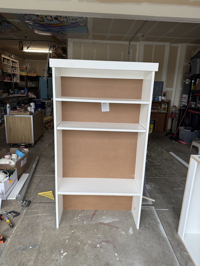 A white-painted bookshelf standing in the workshop, open shelves with a hardboard back.