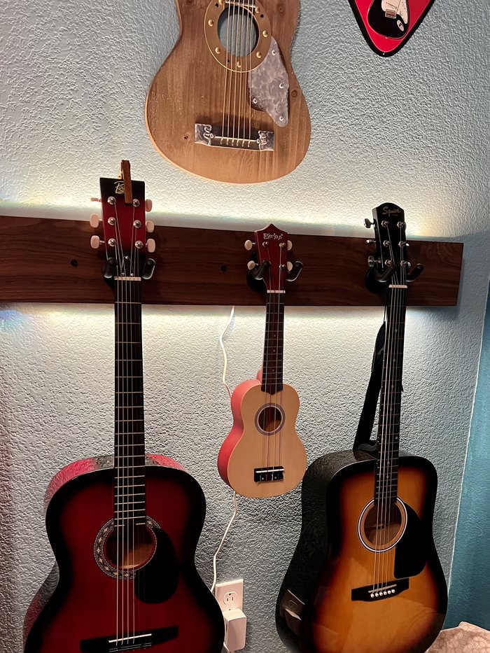 A wall-mounted wooden guitar rack holding two guitars and a ukulele, in daylight.