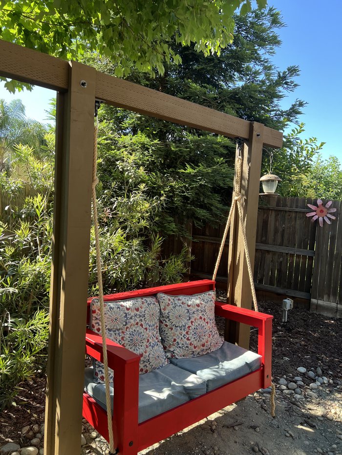 A red-painted porch swing hanging from a timber A-frame in a garden, with cushions on the seat.